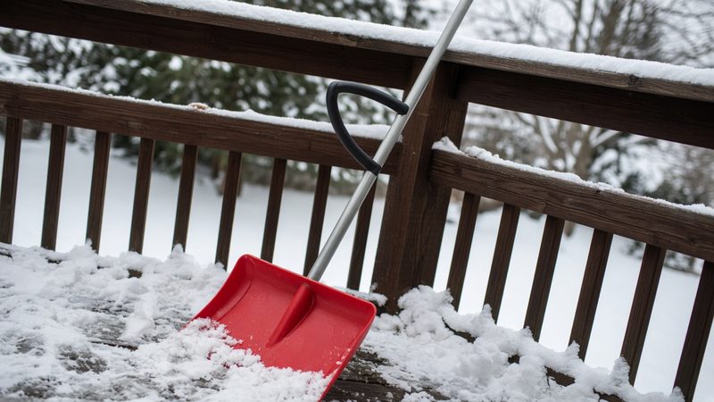 A red snow shovel with a second curved handle leaning on a snowy deck railing, in Syracuse, New York
